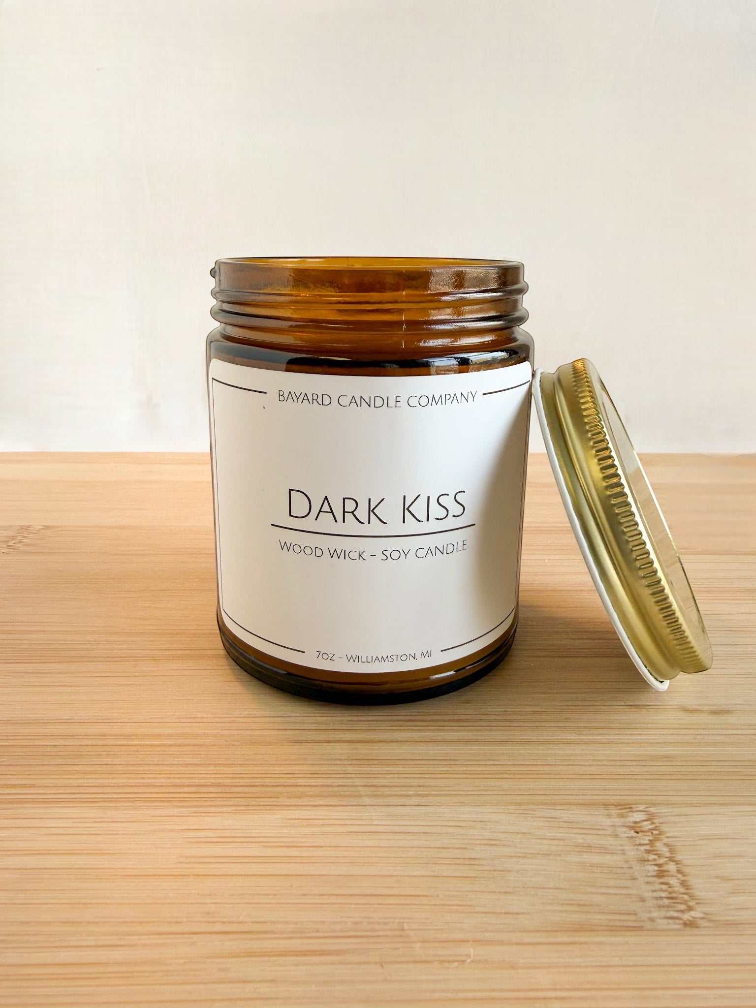 Dark Kiss wood wick soy candle with lid off in amber jar, showing candle detail and gold lid on wood surface