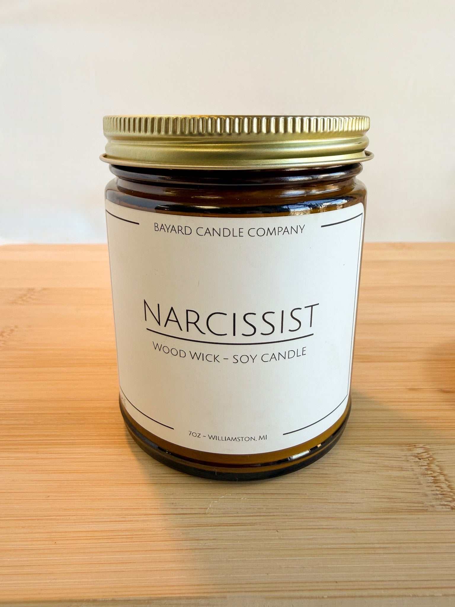 Narcissist wood wick soy candle in amber glass jar with gold lid on wood surface, clean minimalist style