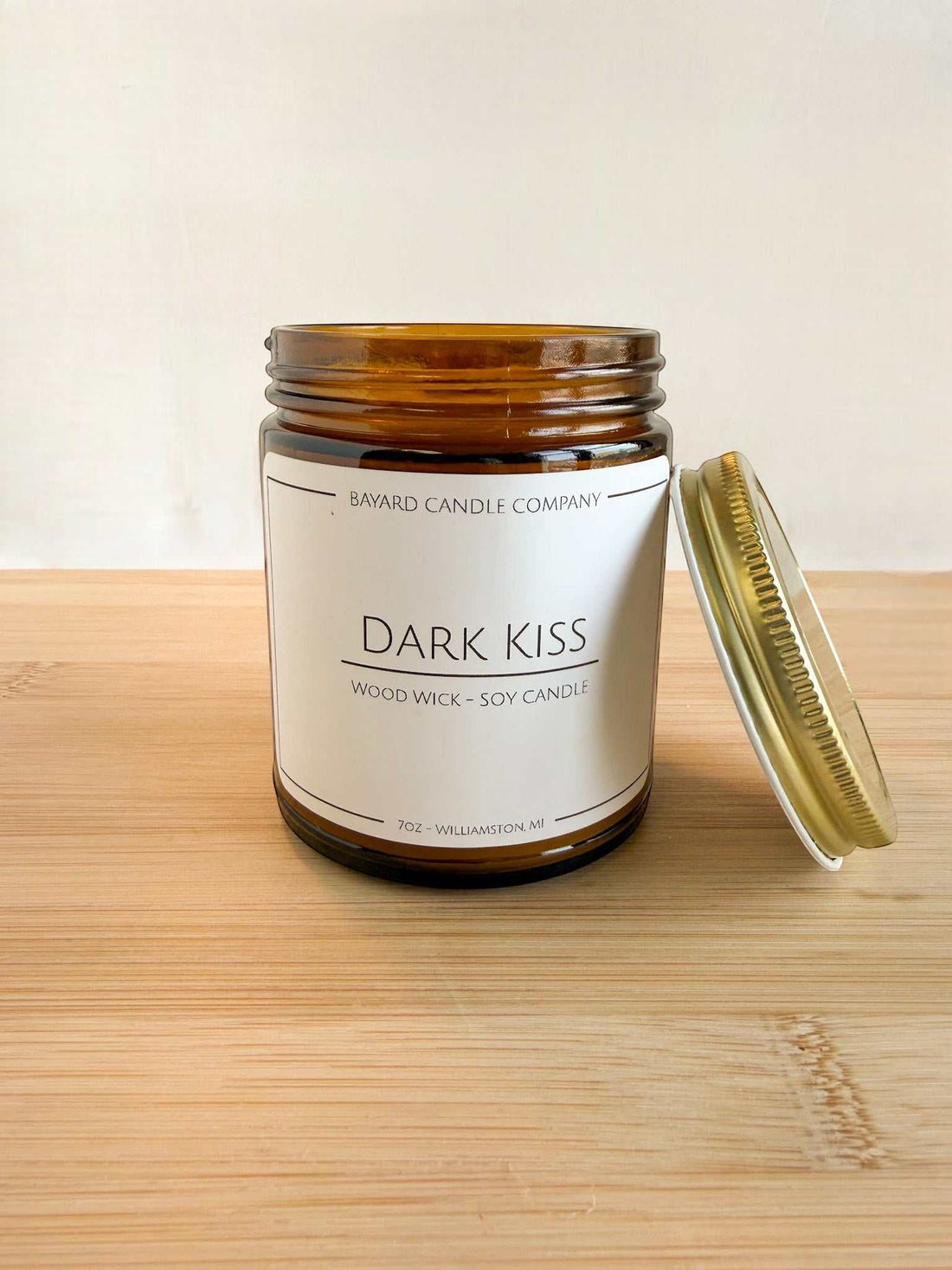 Dark Kiss wood wick soy candle with lid off in amber jar, showing candle detail and gold lid on wood surface
