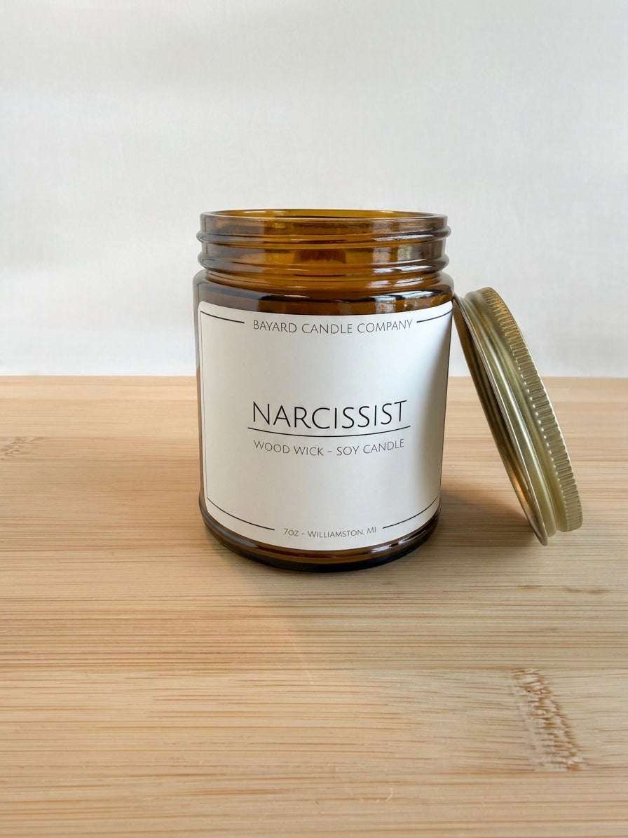 Narcissist wood wick soy candle with lid off  leaning on the side in amber jar