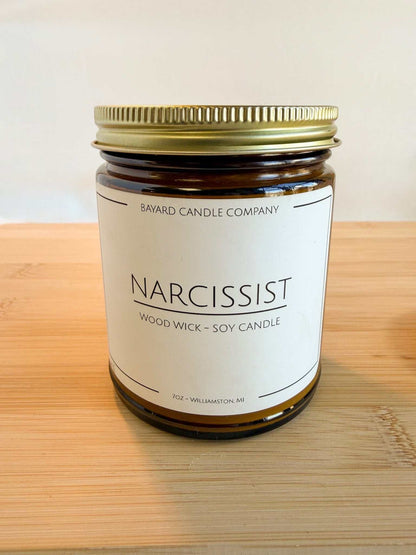 Narcissist wood wick soy candle in amber glass jar with gold lid on wood surface, clean minimalist style