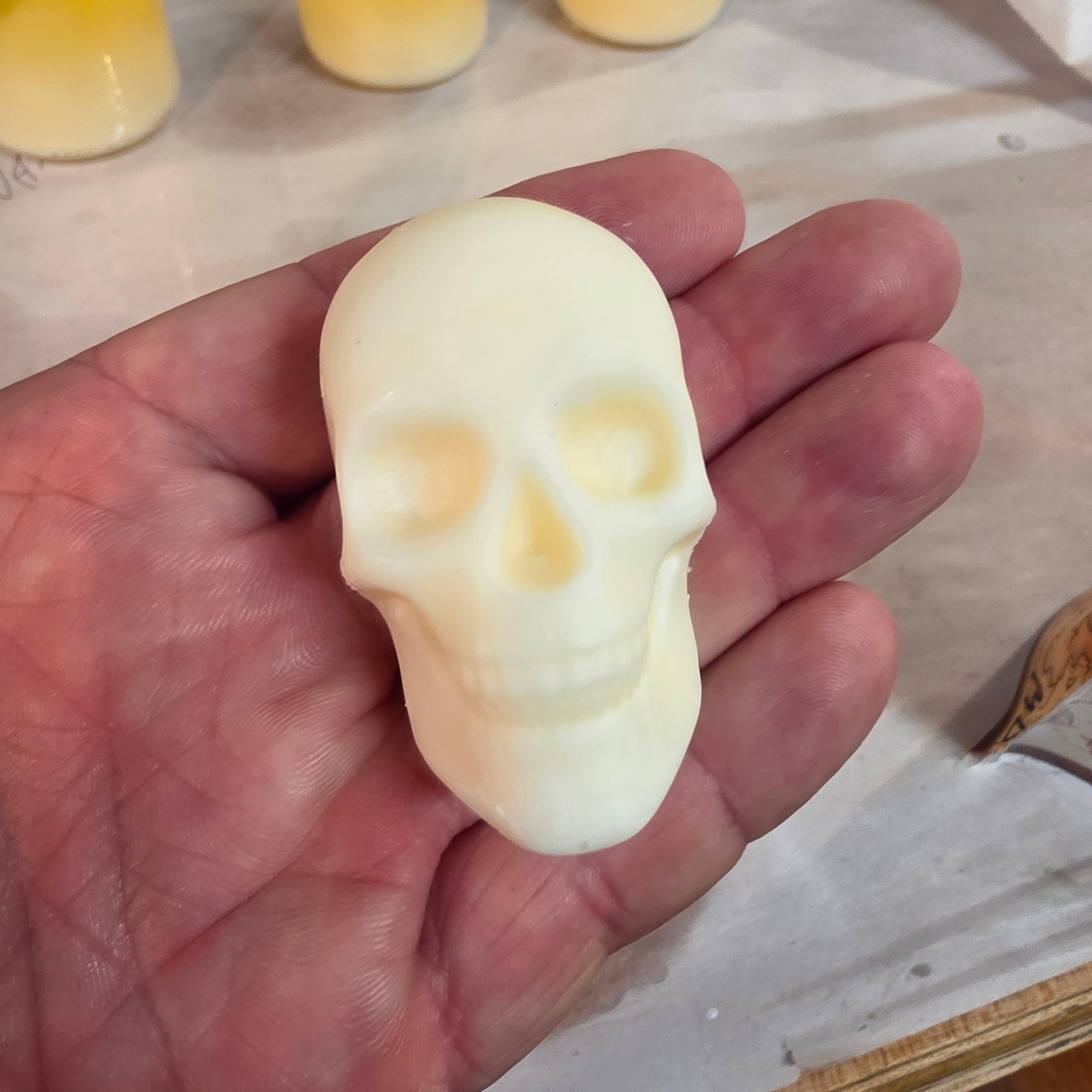 A hand holding a small, off-white skull-shaped wax melt, against a light tabletop background with blurred yellow items in the distance.