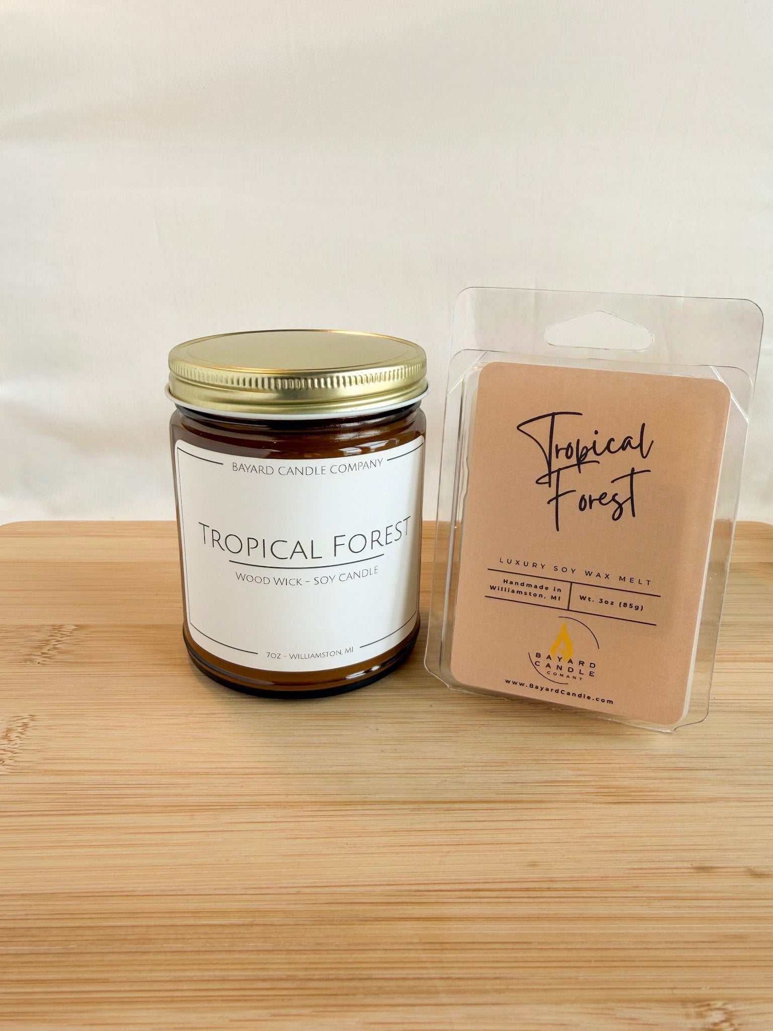 Tropical Forest wood wick soy candle in amber glass jar with gold lid on wood surface next to Tropical Forest Wax Melt, clean background